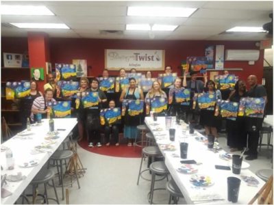 About 20 or so students hold up their version of Van Gogh’s Starry Night in the Painting with a Twist studio in Arlington, Texas. Long tables holding painting supplies are in the foreground.