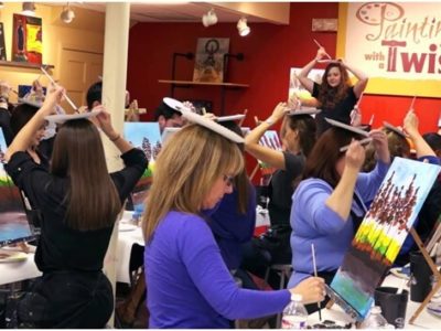 A woman standing in front of a studio class holds a paper plate palette and paintbrush over her head as her students do the same. A Painting with a Twist banner is in the background.