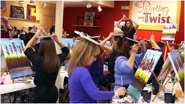 A woman standing in front of a studio class holds a paper plate palette and paintbrush over her head as her students do the same. A Painting with a Twist banner is in the background.