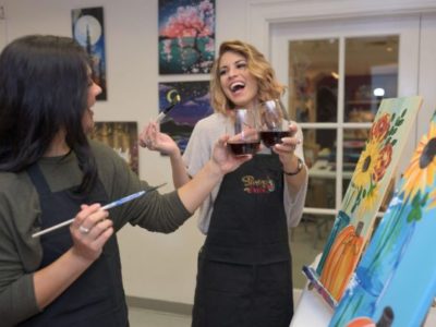 Two women stand at easels with paint brushes, laughing and talking, as they clink their wine glasses together in celebration.