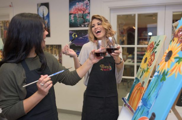 Two women stand at easels with paint brushes, laughing and talking, as they clink their wine glasses together in celebration.