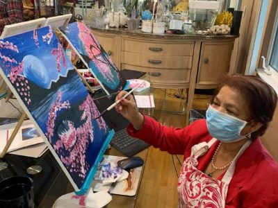 A woman in a surgical face mask and a red shirt and apron paints a canvas of moonlit cherry blossoms in a Painting with a Twist studio.